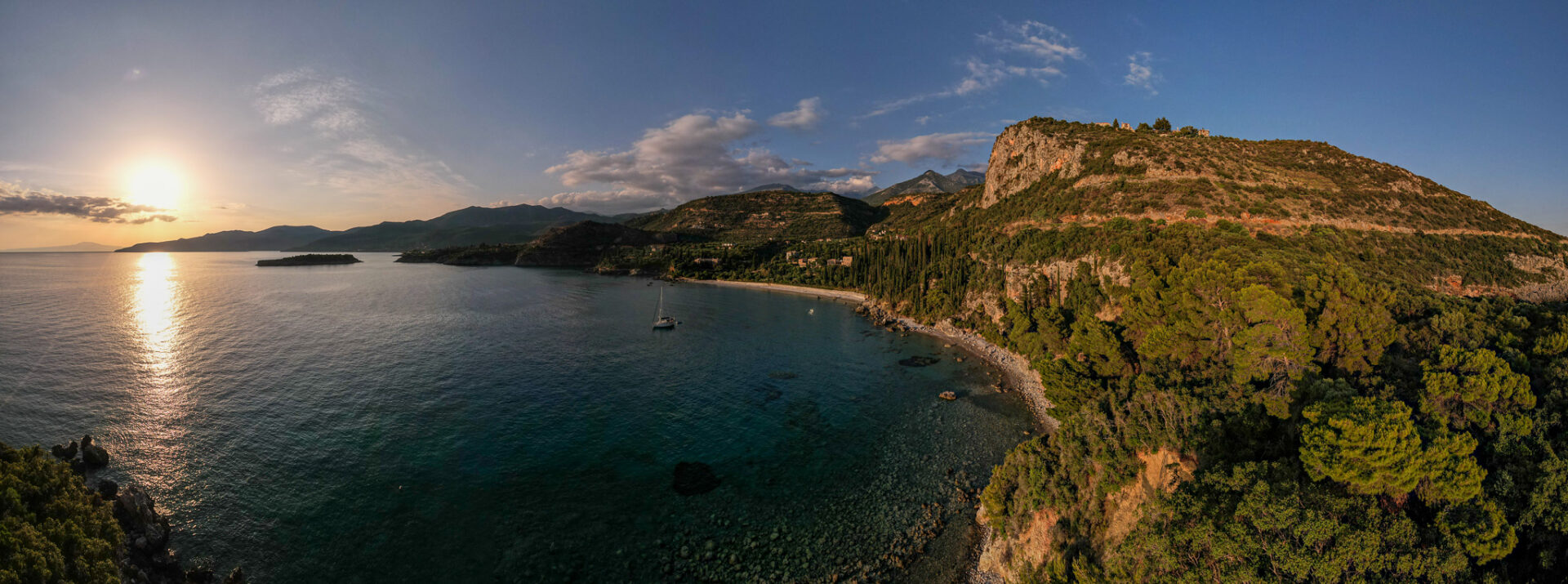 Sunset view over Kardamili's coastline with a sailboat on the ocean, rugged mountains in the background, and lush greenery along the shore.
